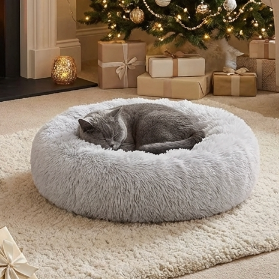Gray cat sleeping in a light gray fluffy donut bed in a warm Christmas room with a decorated tree and wrapped presents.