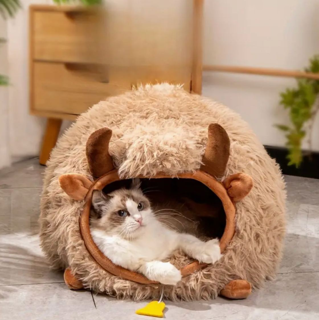 Cat lying inside a fluffy pet bed with animal ears on a wooden floor.
