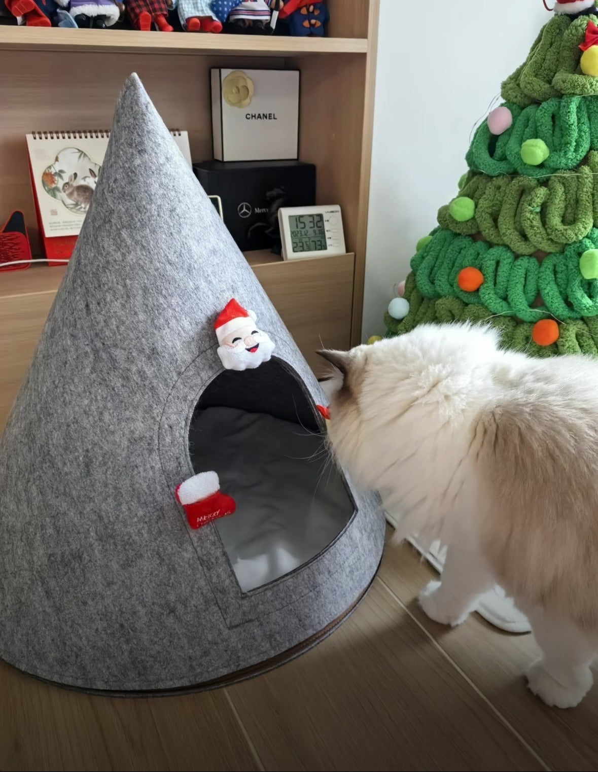 Fluffy white cat inspecting a gray conical cat bed with Santa Claus decorations, placed next to a festive Christmas setup.