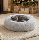 Gray cat sleeping in a light gray fluffy donut bed in a warm Christmas room with a decorated tree and wrapped presents.