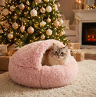Cat sitting inside a pink fluffy cozy cocoon bed in a warm Christmas living room with a decorated tree and fireplace